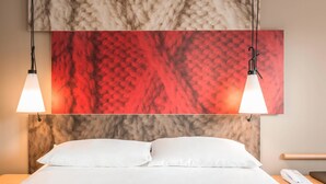 Una habitación de hotel con una cama, almohadas blancas, una mesita de noche con teléfono y dos lámparas colgantes.