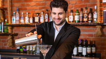 Un cantinero sirviendo una bebida en una copa de martini en un bar con un fondo de pared de ladrillo y estantes llenos de botellas de licor.