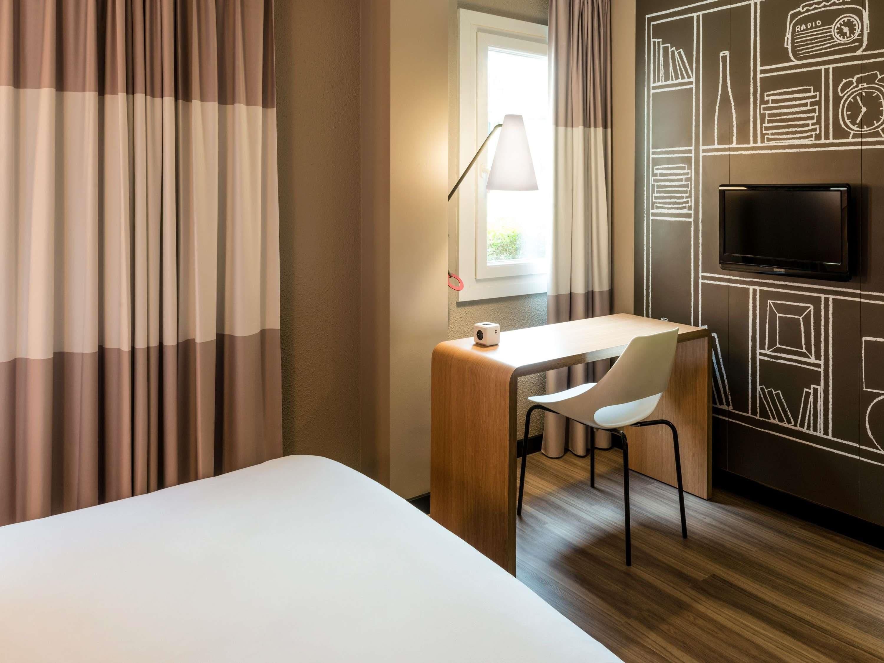 A hotel room with a bed, a desk with a chair, a lamp, a TV, and a window with curtains.