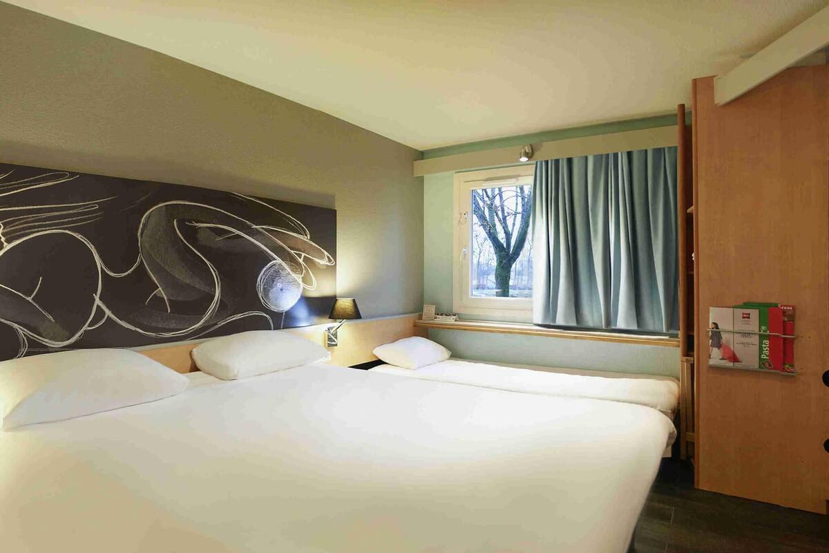 Una habitación de hotel con una cama grande, una ventana con cortinas, una lámpara fijada a la pared y un estante con objetos.