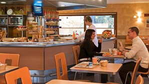 Ein Café mit einer Theke, an der Gebäck und eine Kaffeemaschine präsentiert werden. Es gibt Tische und Stühle für die Gäste.