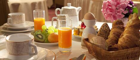 Um cenário de café da manhã com uma mesa posta para dois, incluindo pratos, xícaras, um jarro de suco de laranja, uma cesta de croissants e um vaso de flores.