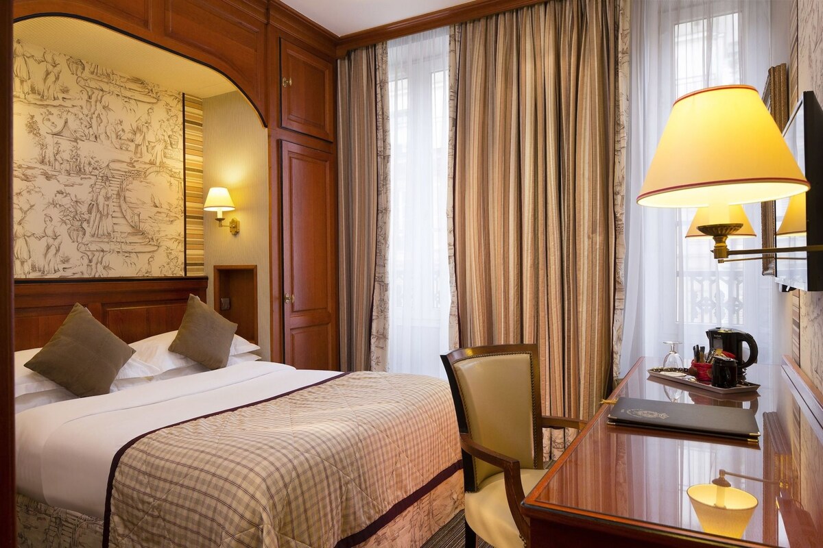 A hotel room with a bed, a desk with a lamp, a chair, and a large window with curtains.