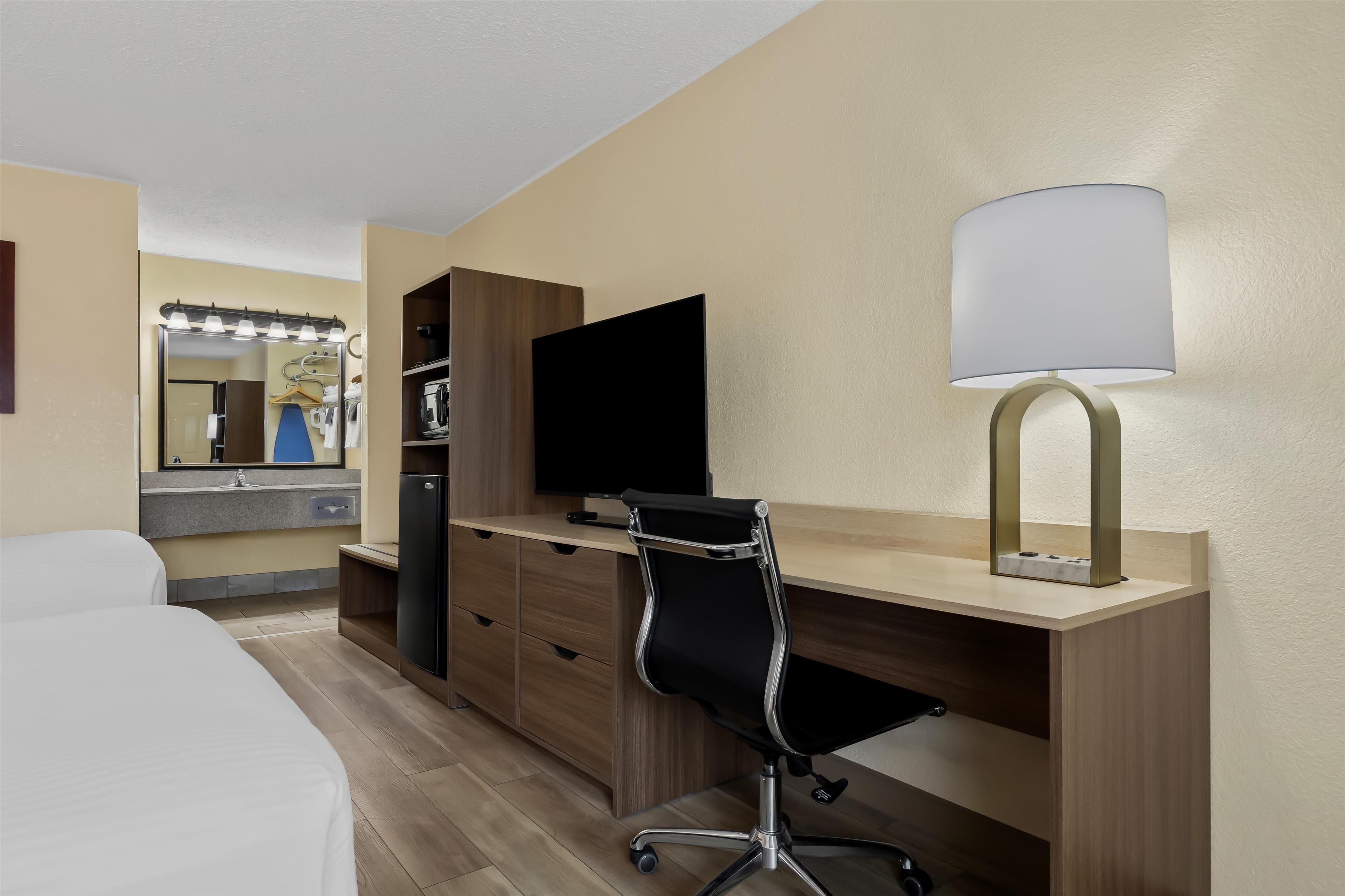 A hotel room with a bed, a desk with a computer, a lamp, a TV, a mirror, and a wardrobe.