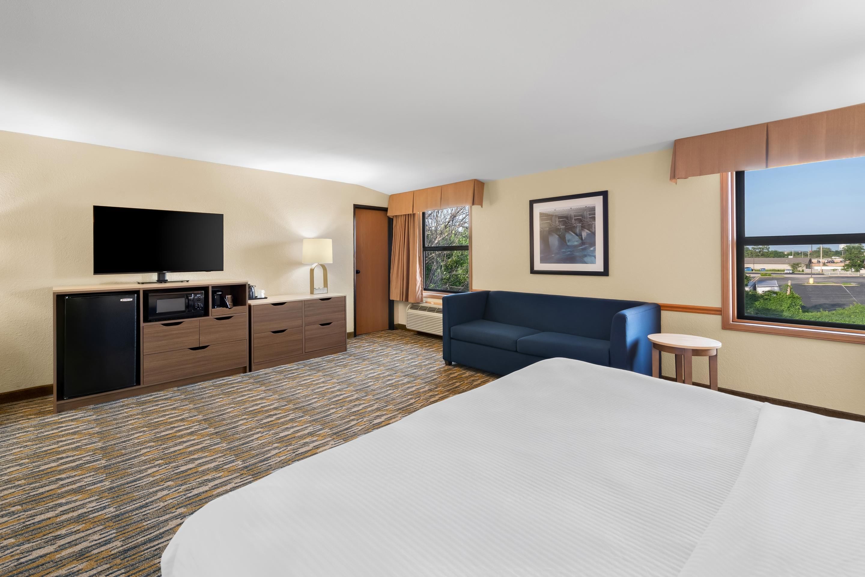 A hotel room with a bed, a television, a desk with a lamp, a sofa, and a window with a view of a parking lot.