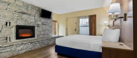 Habitación de hotel con chimenea de piedra, cama con cabecera azul, mesita de noche de madera y televisor montado en la pared.