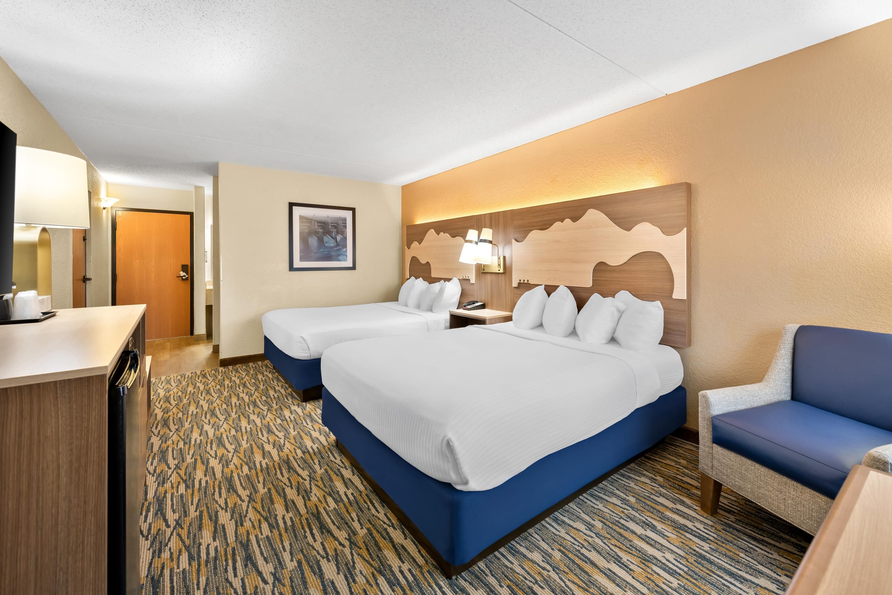 A hotel room with two beds, a desk, a chair, a wall-mounted lamp, and a framed picture.