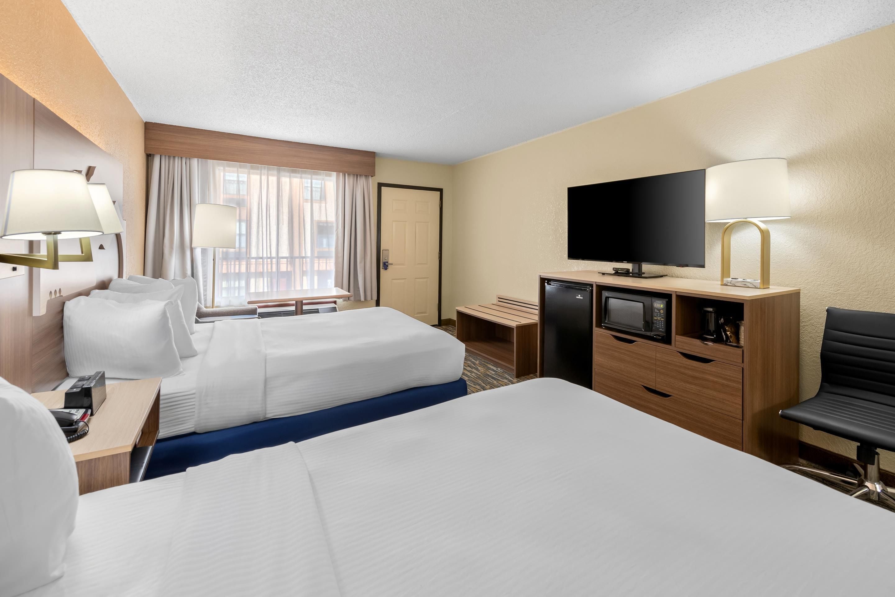 A hotel room with two beds, a desk with a telephone, a flat-screen TV, a minibar, and a seating area with a chair.