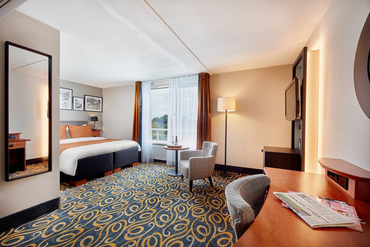A hotel room with a bed, a desk, a chair, a small table, and a window with curtains.