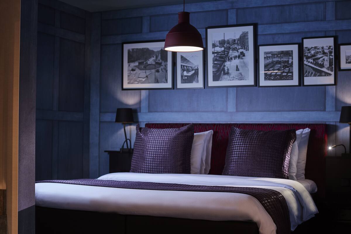 A bedroom with a bed, a hanging red pendant light, and a wall with framed black and white photographs.