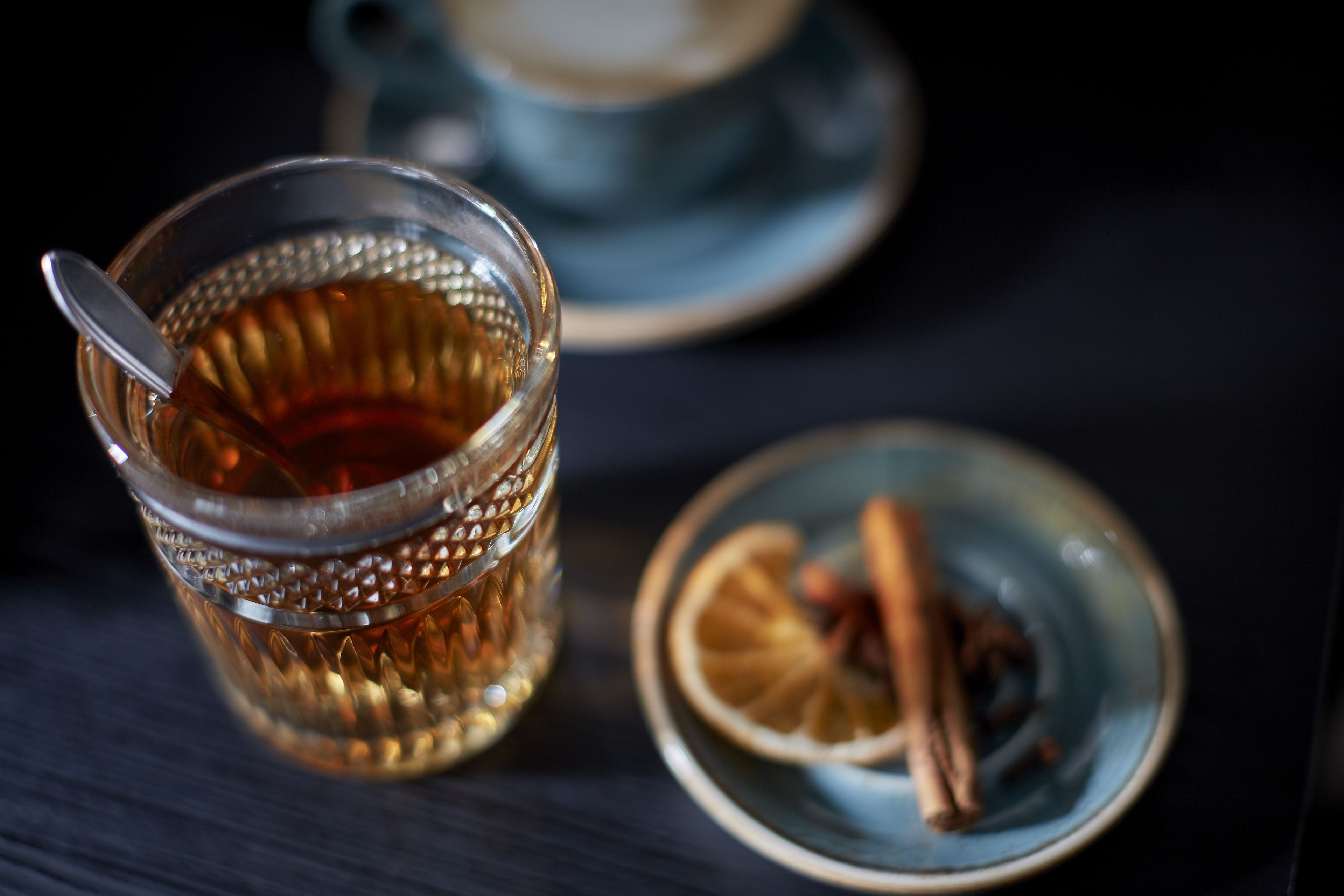 A glass of tea with a spoon, a plate with cinnamon sticks and a slice of lemon, and a blue teapot in the background.