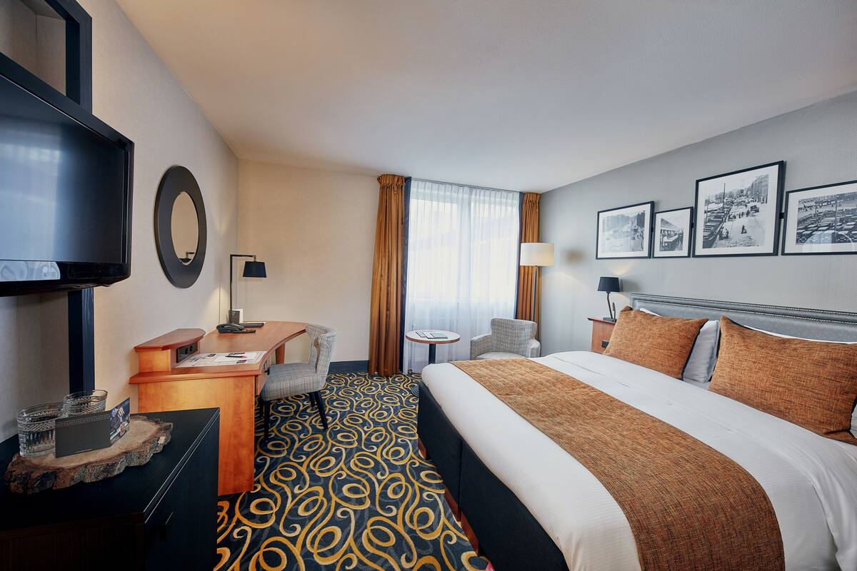 A hotel room with a bed, a desk, a television, a chair, a small table, and a window with curtains.