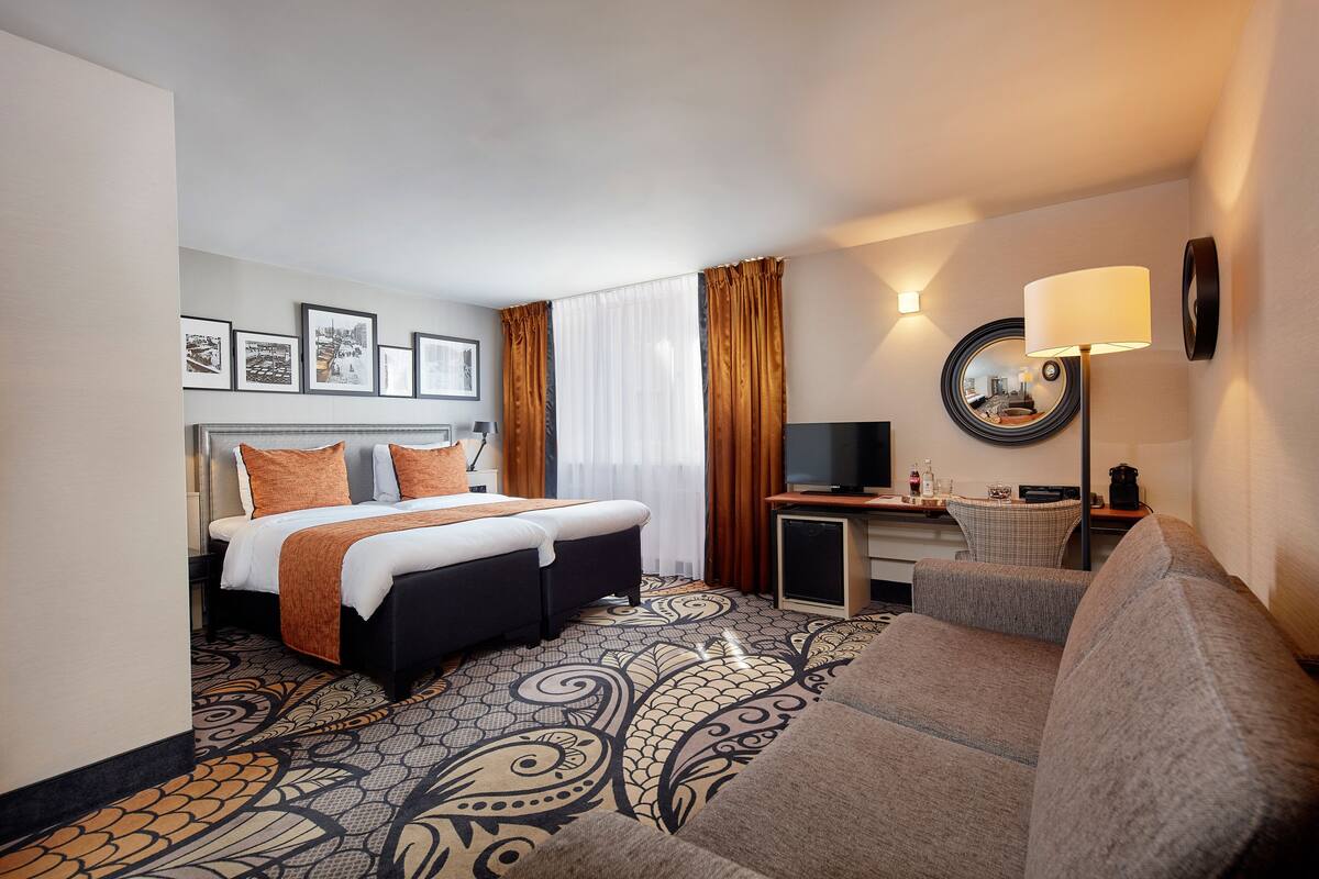 A hotel room with a bed, a sofa, a desk with a TV, and a mirror on the wall.