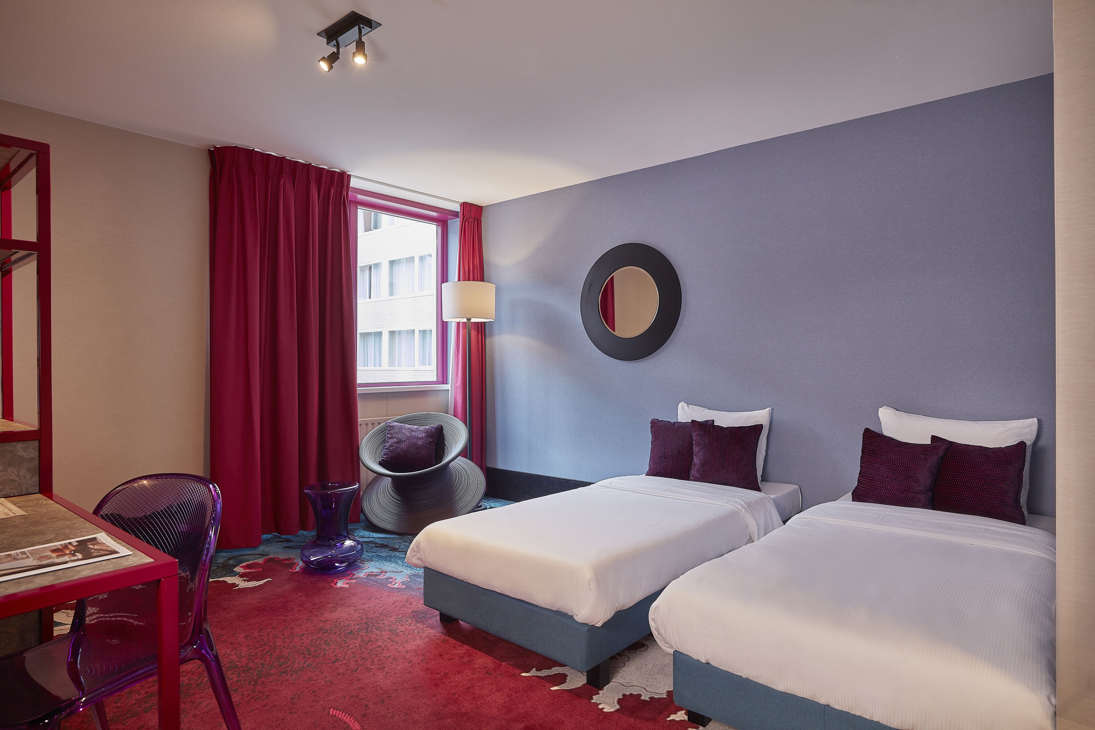 A hotel room with two beds, a desk, a chair, a round mirror, and a window with red curtains.