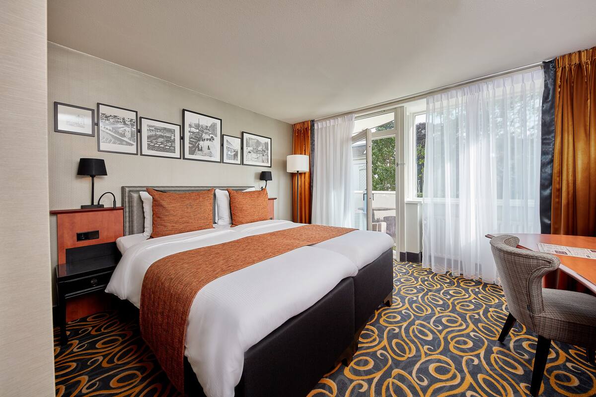 A hotel room with a large bed, a desk, and a chair. There are framed pictures on the wall and a window with curtains.