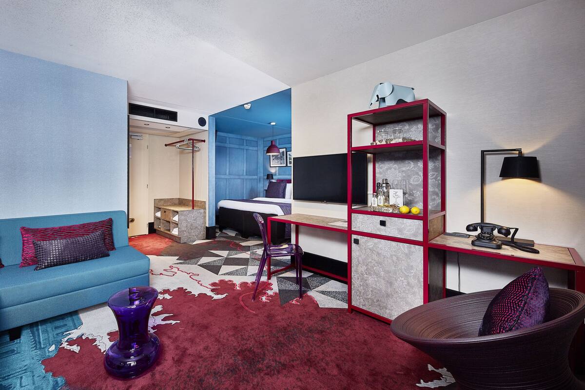 A modern hotel room with a blue sofa, a red and white patterned rug, a purple vase, a desk with a lamp, and a chair.