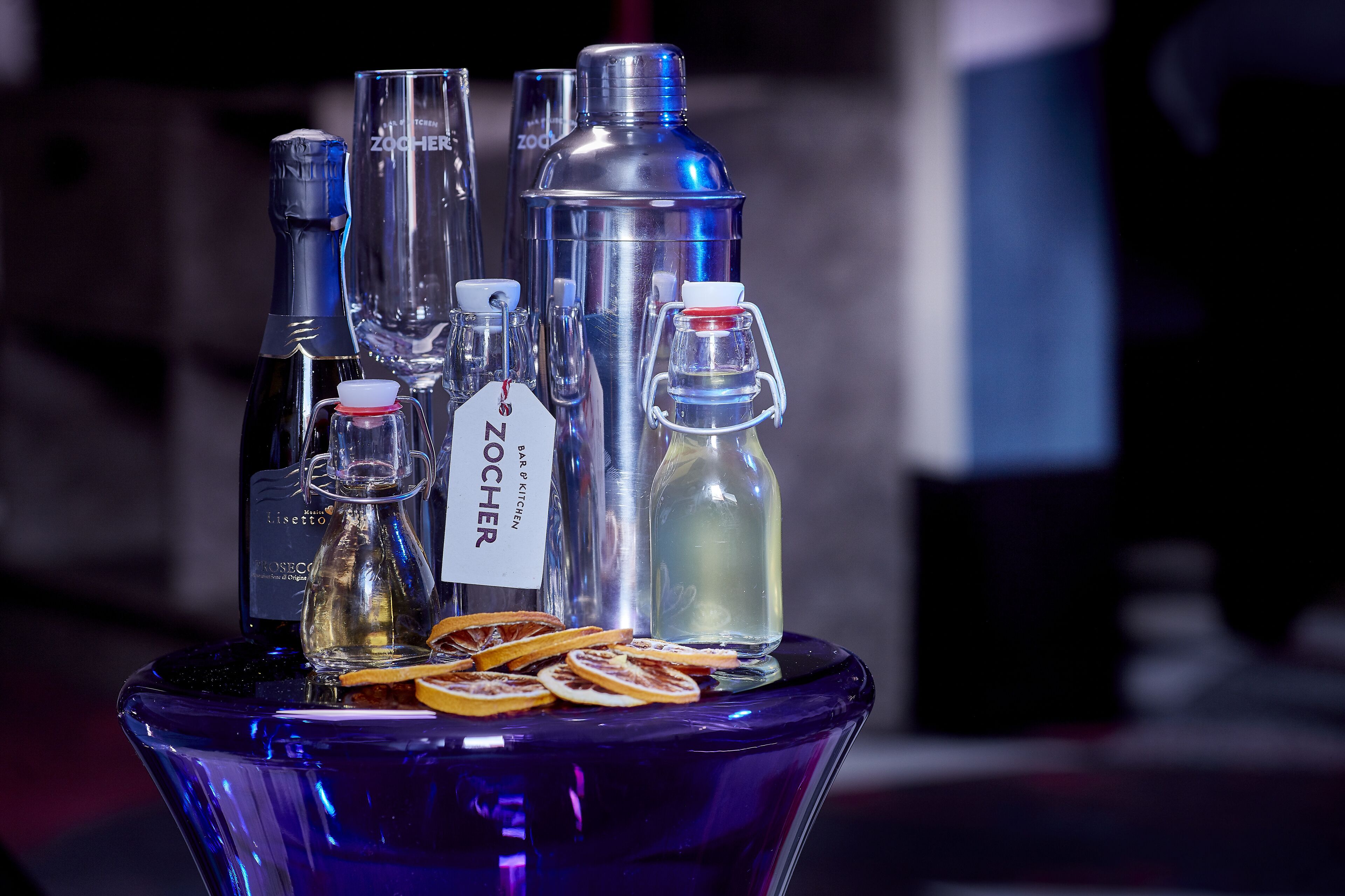 Une sélection de bouteilles et de verres est disposée sur une table bleue, et l’une des bouteilles porte l’étiquette « ZOOCHER ».