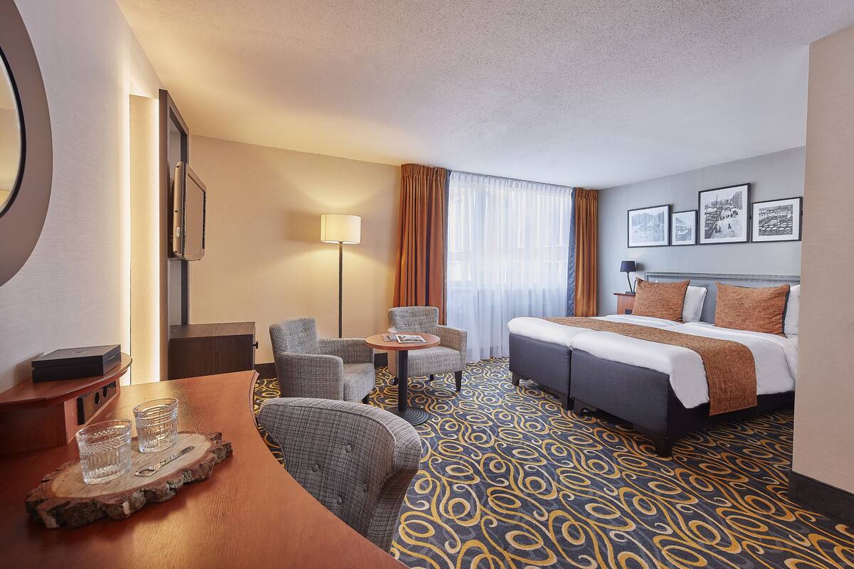 A hotel room with a bed, two armchairs, a small table with glasses, a lamp, and a wall with framed pictures.