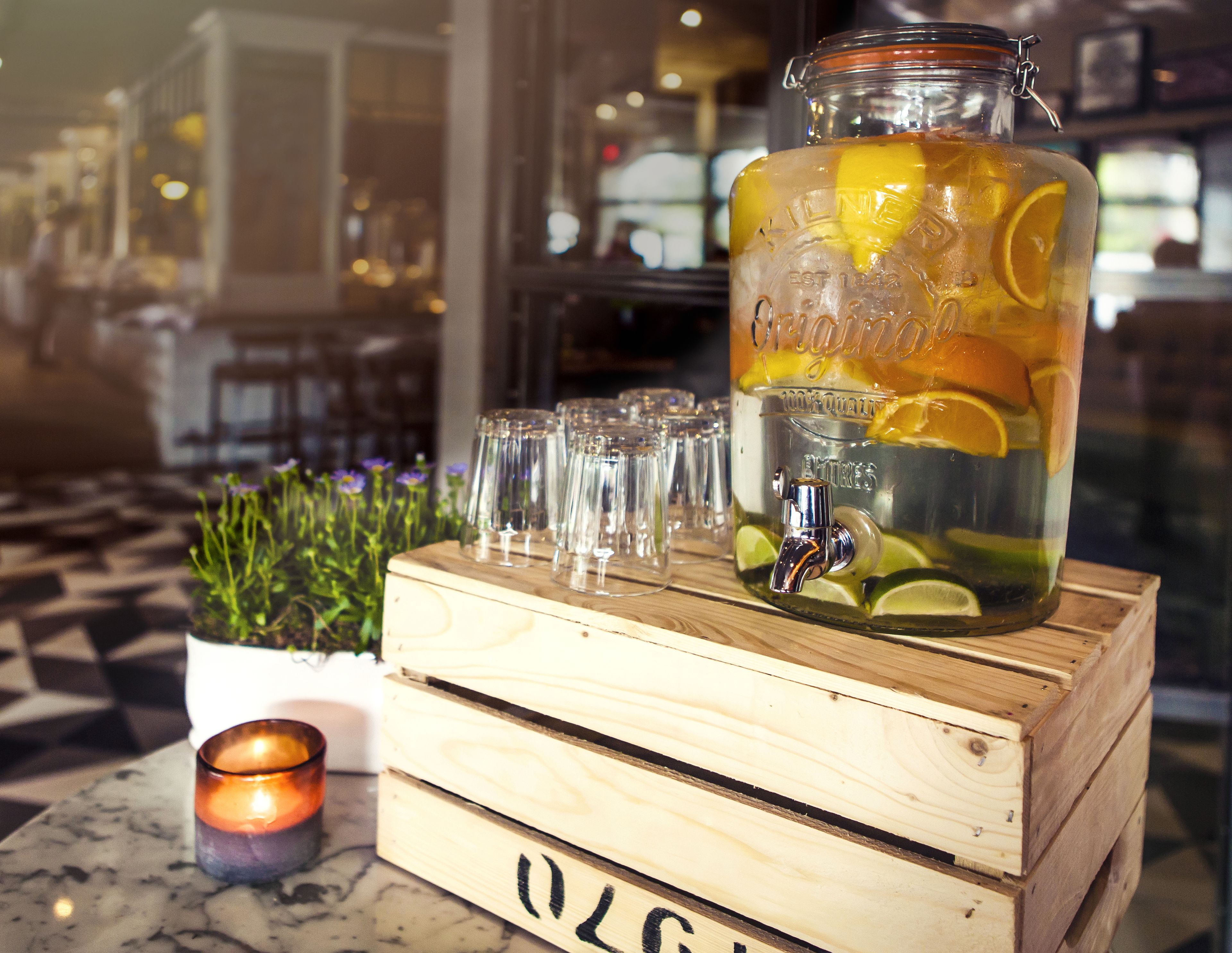 Une caisse en bois contenant un bocal en verre rempli d’une boisson, des verres et une bougie, le tout posé sur une table.
