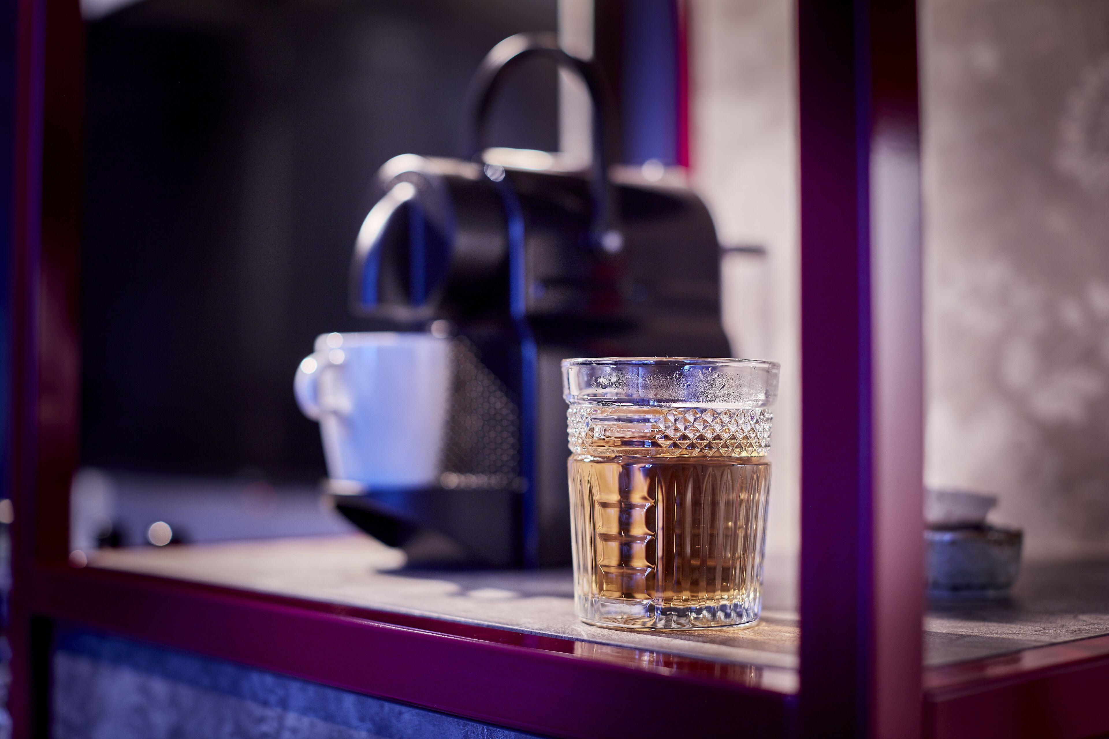 A glass of amber liquid on a shelf with a coffee maker in the background.