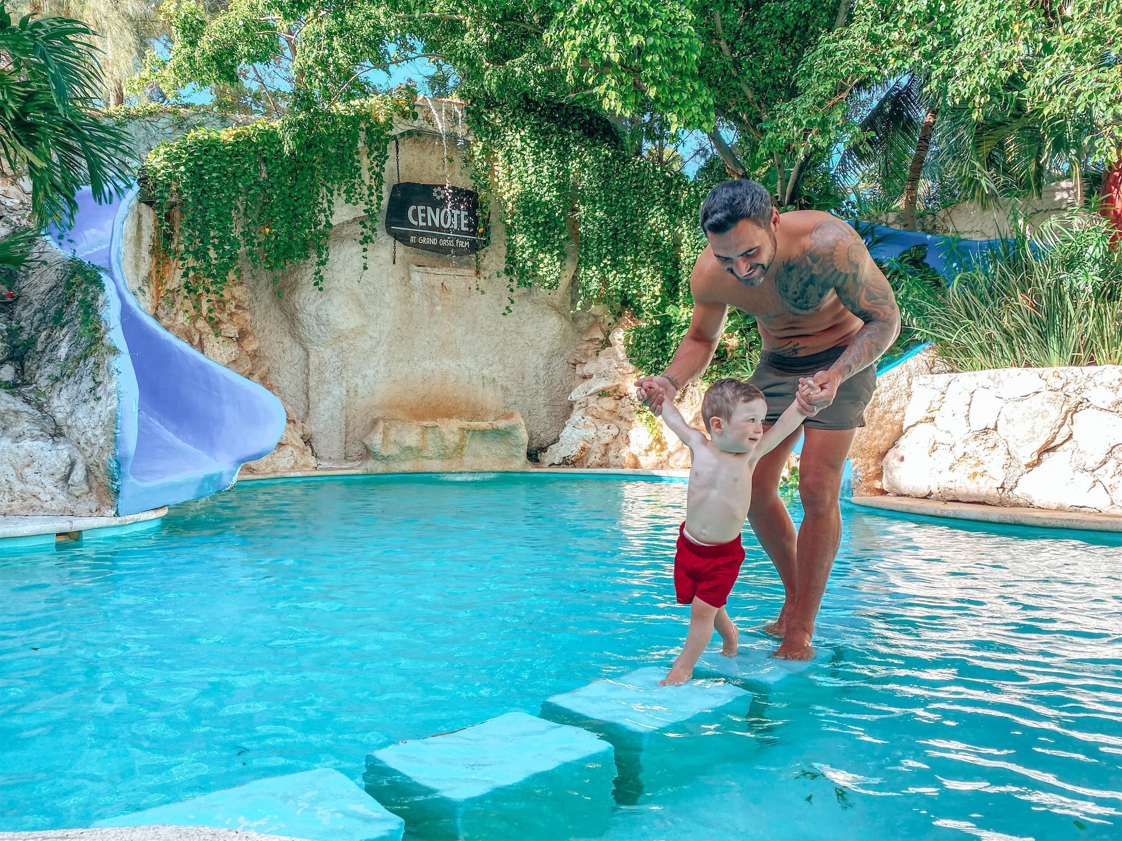 Een man en een kind in een zwembad met een glijbaan en een bord waarop staat 'CENOTE IT GRAND OASIS FARM'.