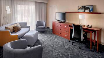 A hotel room with a grey sofa, a desk with a computer, a television, a lamp, and a door.