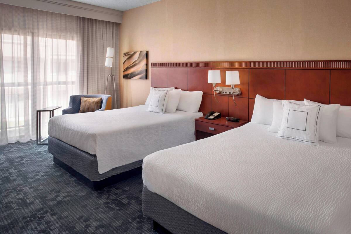 Habitación de hotel con dos camas, una mesita de noche con lámpara, una silla y un ventanal con cortinas traslúcidas.