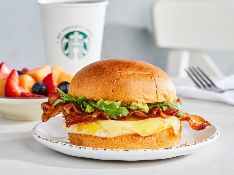 A breakfast sandwich with bacon, lettuce, and cheese on a bun, accompanied by a cup and a bowl of fruit.
