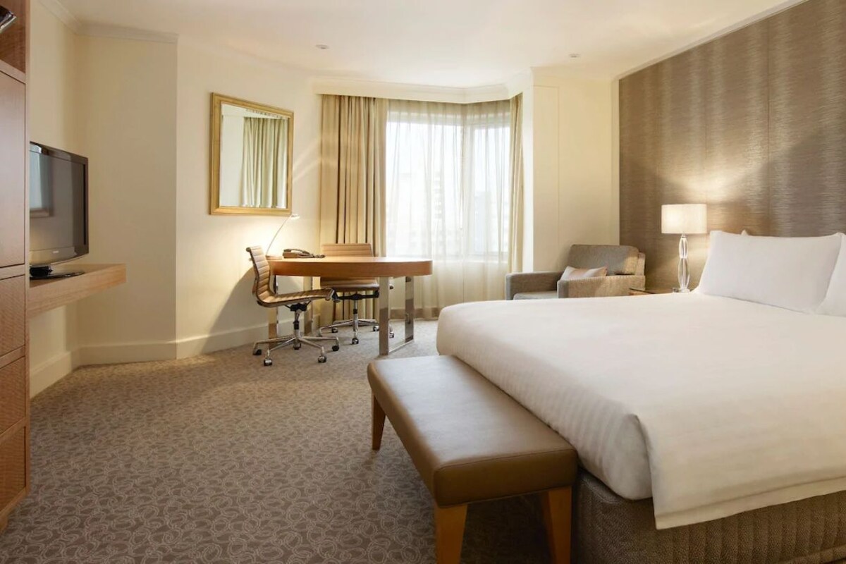 Ein Hotelzimmer mit einem großen Bett, einem Schreibtisch mit Stuhl, einem Fernseher und einem Fenster mit Vorhängen.
