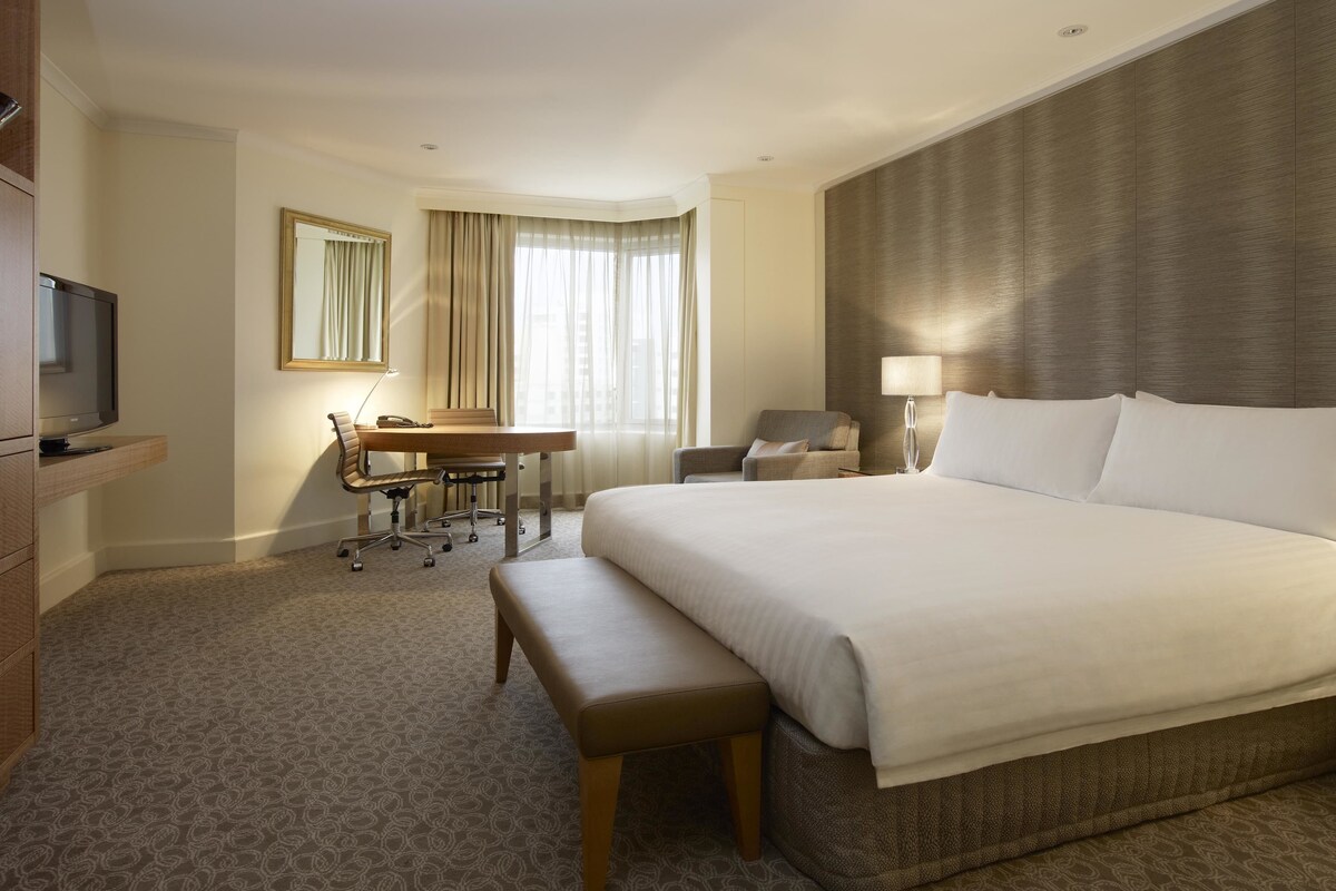 Ein Hotelzimmer mit einem großen Bett, einem Schreibtisch mit Stuhl, einem Fernseher und einem Fenster mit Vorhängen.