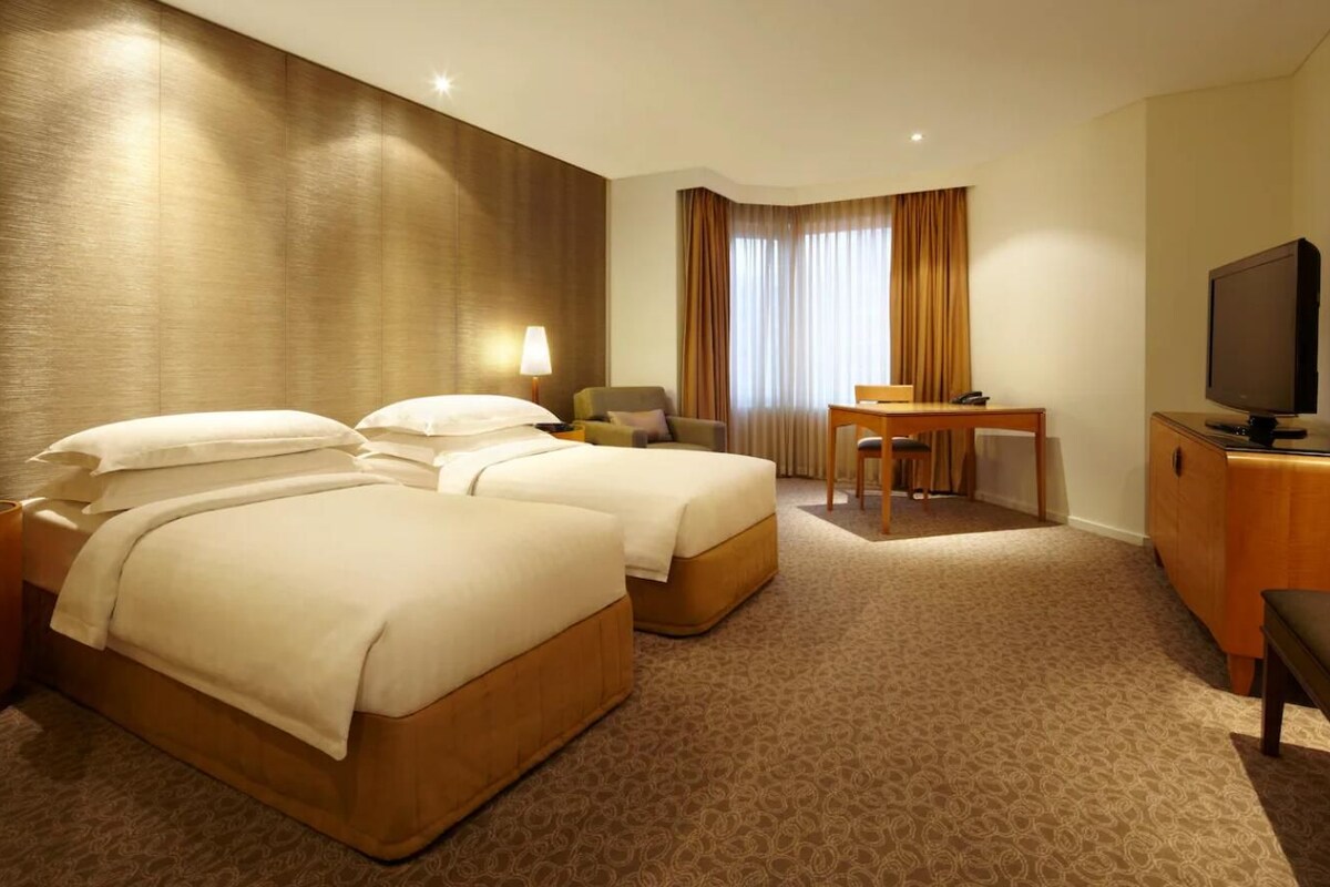 Ein Hotelzimmer mit zwei Betten, einem Schreibtisch, einem Stuhl, einem Fernseher und einem Fenster mit Vorhängen.