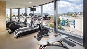 Ein Fitnessstudio mit Laufbändern, Crosstrainern und einer Bank, ausgestattet mit großen Fenstern und Blick auf die Stadt.