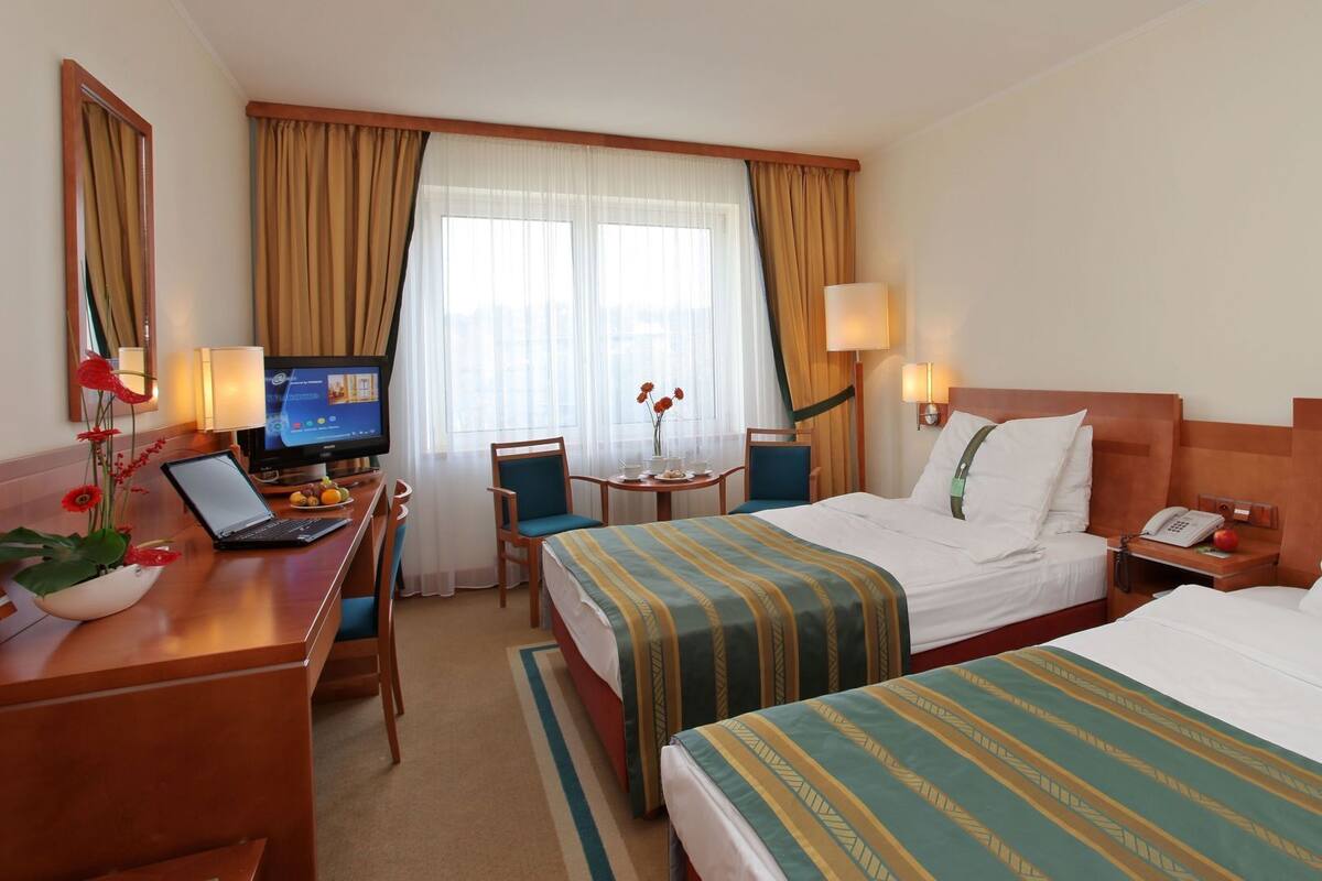 Une chambre d’hôtel avec deux lits, un bureau avec un ordinateur portable, une télévision et une petite table sur laquelle est posé un vase de fleurs.
