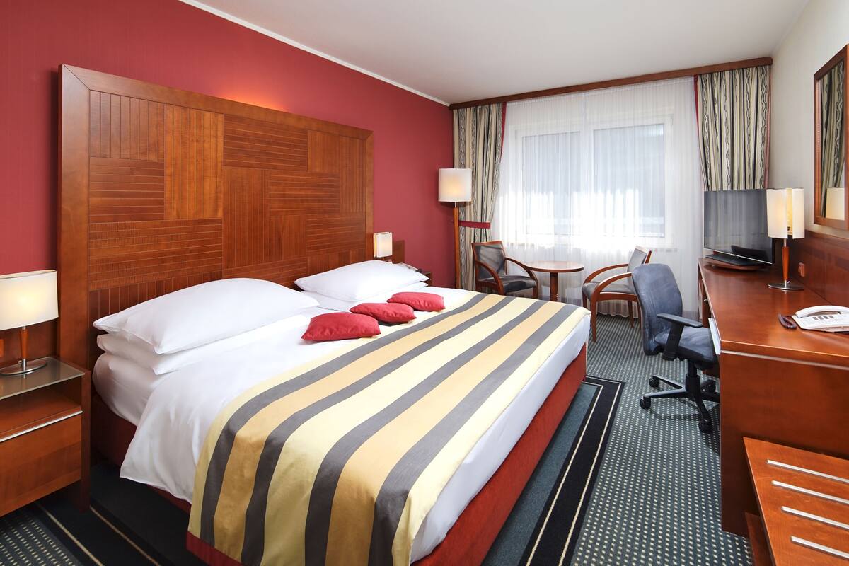 Une chambre d’hôtel avec un grand lit, deux tables de chevet, un bureau avec une chaise, une télévision et une fenêtre avec des rideaux.