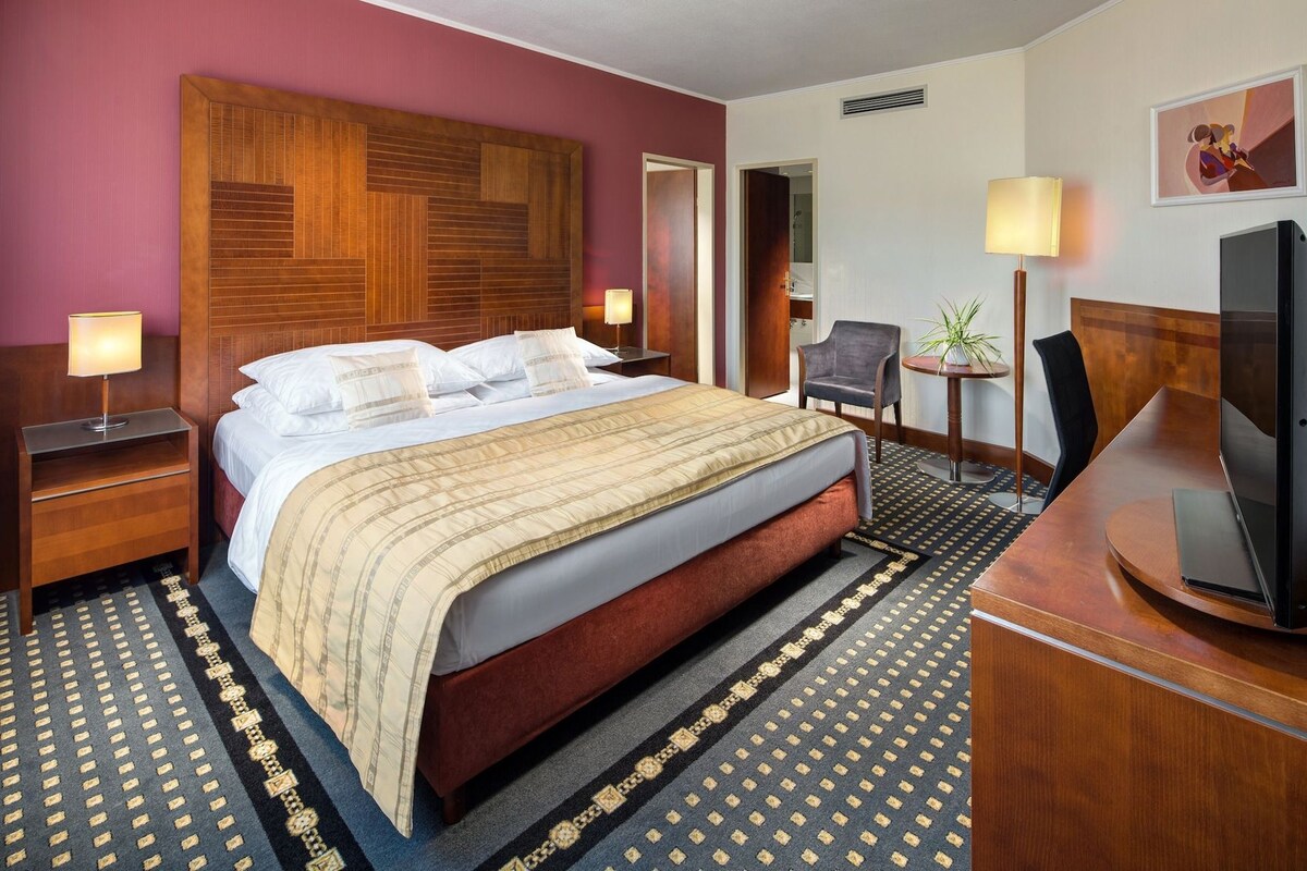 Ein Hotelzimmer mit einem großen Bett, einem Schreibtisch, einem Sessel, einem Fernseher und einem Gemälde an der Wand.
