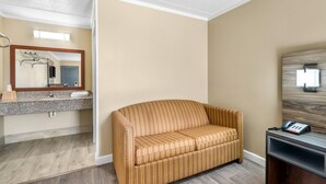 Une chambre d’hôtel avec un mur de couleur beige, un plancher en bois, un canapé, un bureau avec un téléphone et un miroir au-dessus du lavabo.