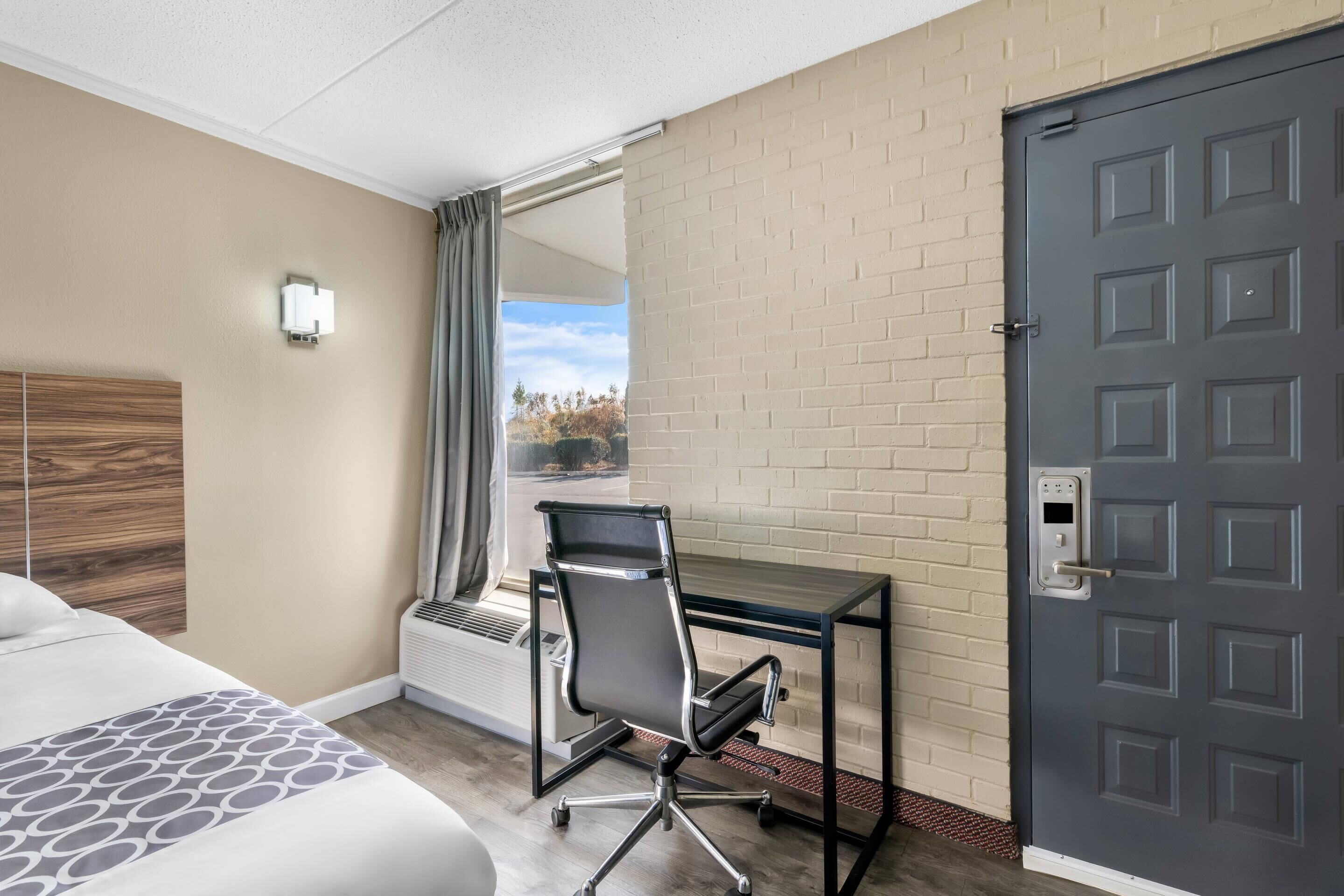 A hotel room with a bed, a desk, a chair, a window with curtains, and a door.