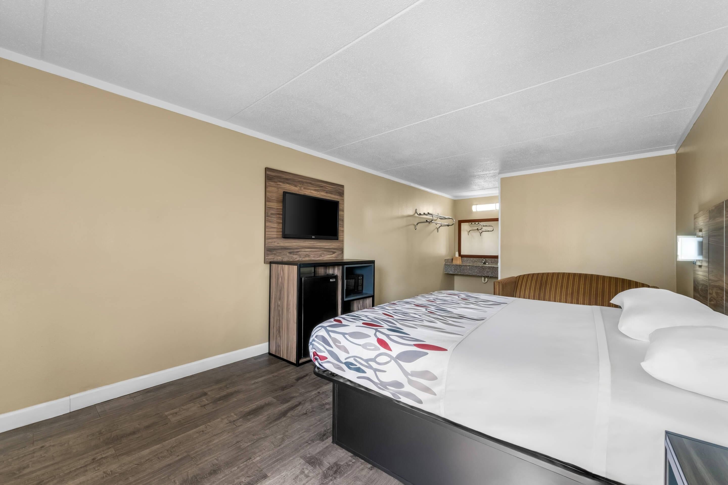 A modern hotel room with a bed, a television mounted on a wooden panel, a small desk with a computer, and a mirror above the bed.