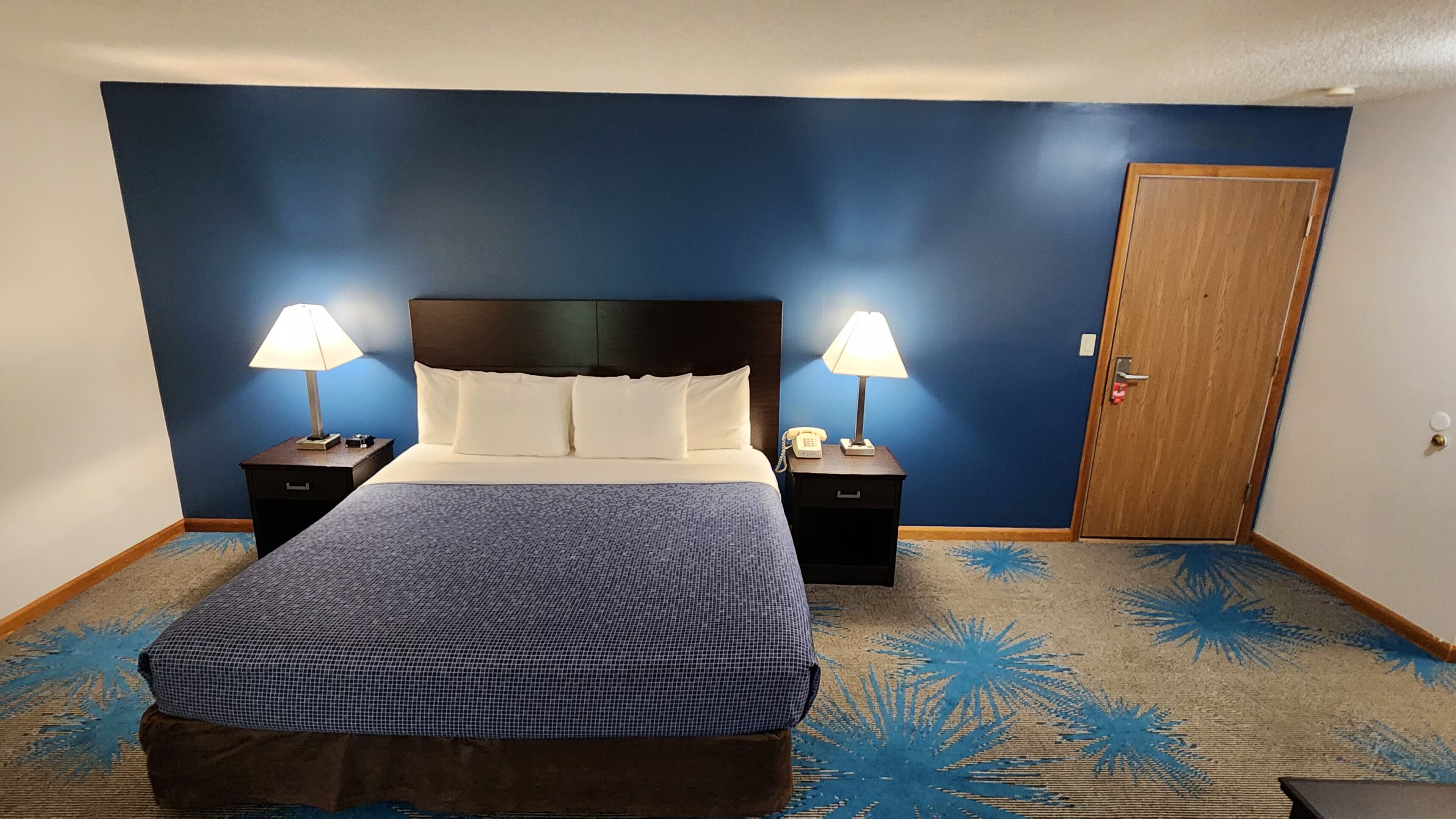 Quarto de hotel com uma cama grande, duas mesas de cabeceira com abajures, uma porta de madeira e um tapete azul com motivos de palmeiras.