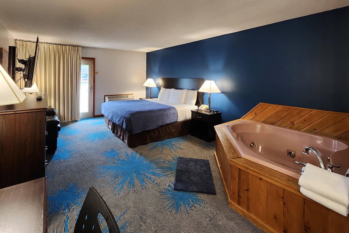 A hotel room with a bed, two bedside tables with lamps, a wooden headboard, a large bathtub, and a blue patterned rug.