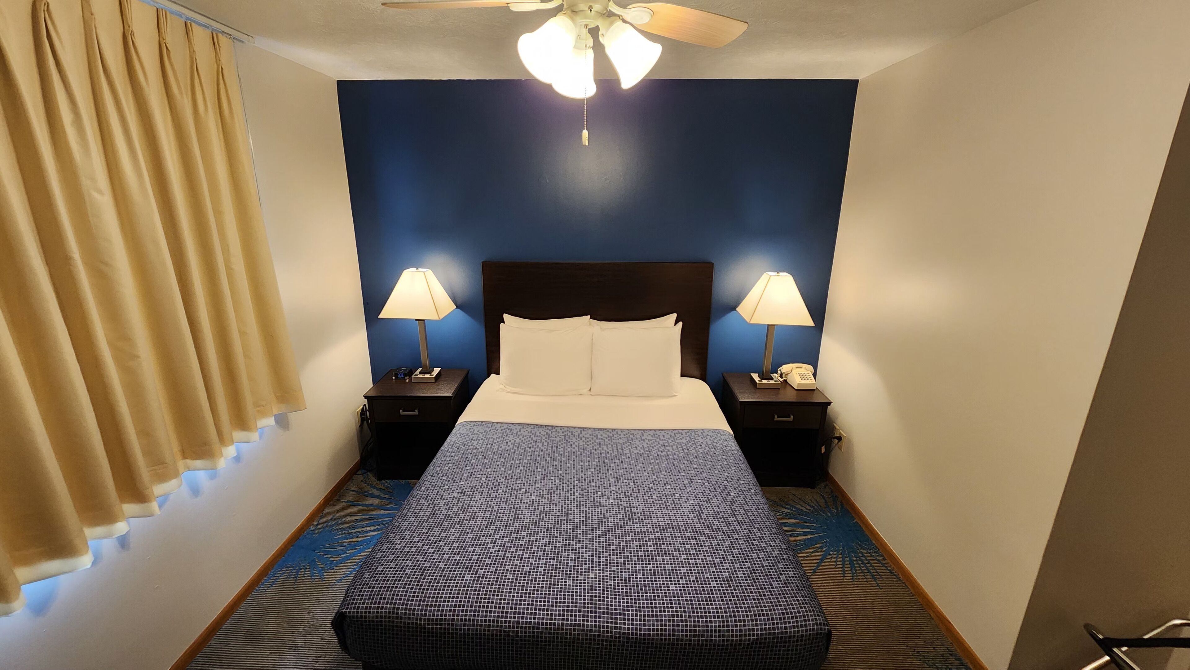 A hotel room with a bed, two bedside tables with lamps, a ceiling fan, and a patterned rug.