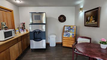 Cocina compacta con refrigerador de acero inoxidable, microondas, armarios de madera, basurero blanco, un tablón de anuncios, una mesa de comedor con una silla y un cuadro en la pared.