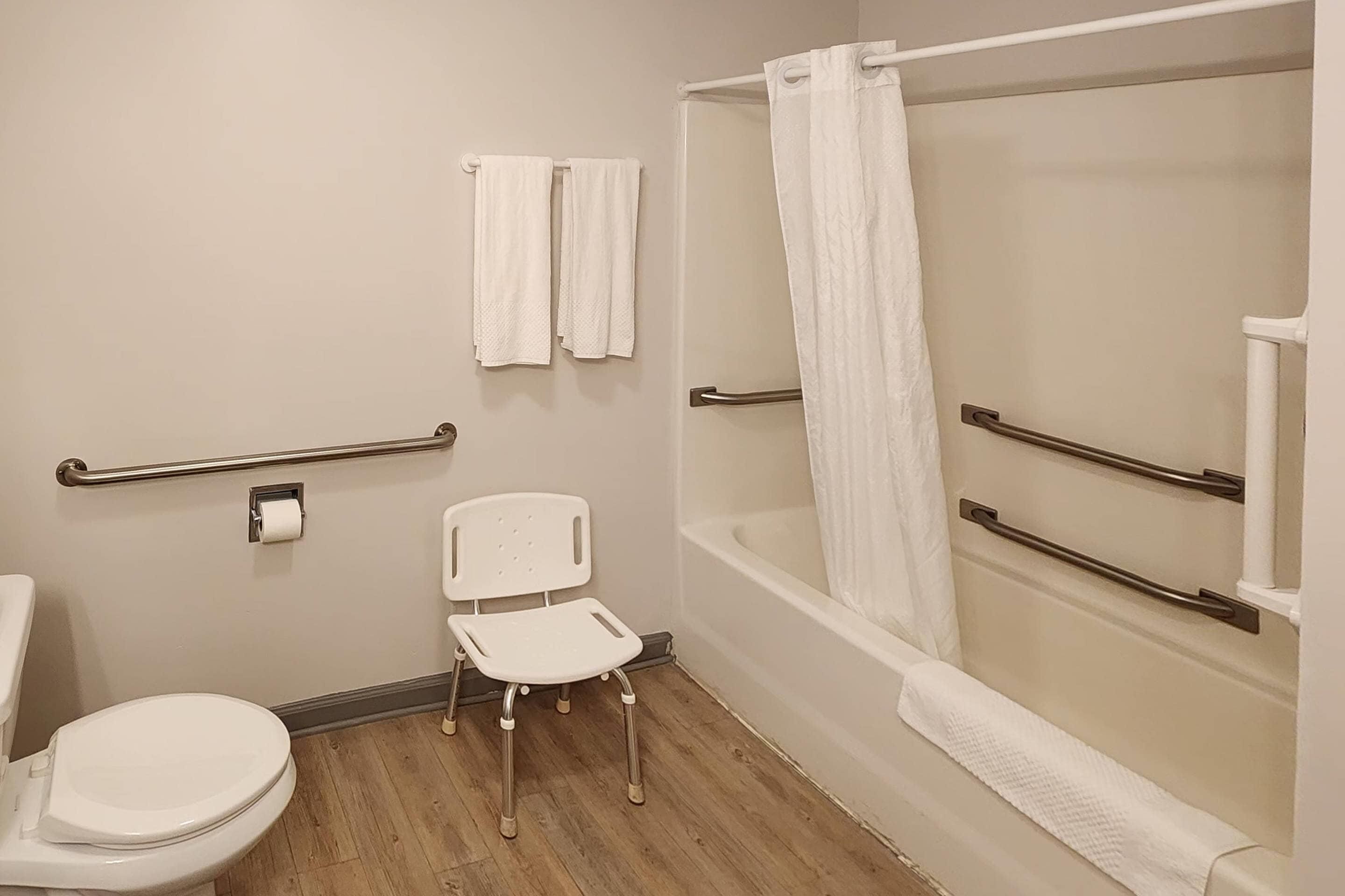 Et badeværelse med toilet, en hvid stol, badekar med bruseforhæng og gelændere.