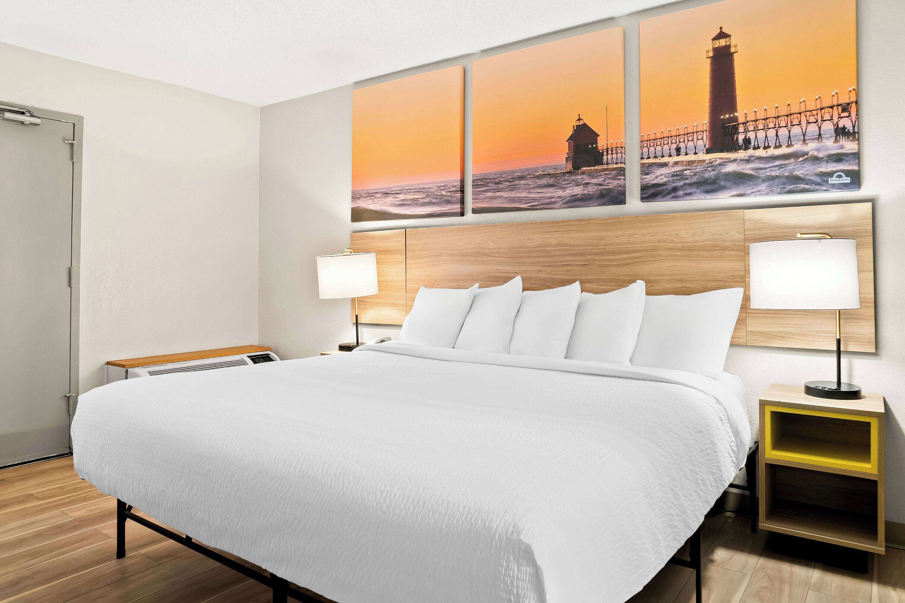 A hotel room with a large bed, two bedside tables, and a wall-mounted painting of a lighthouse at sunset.