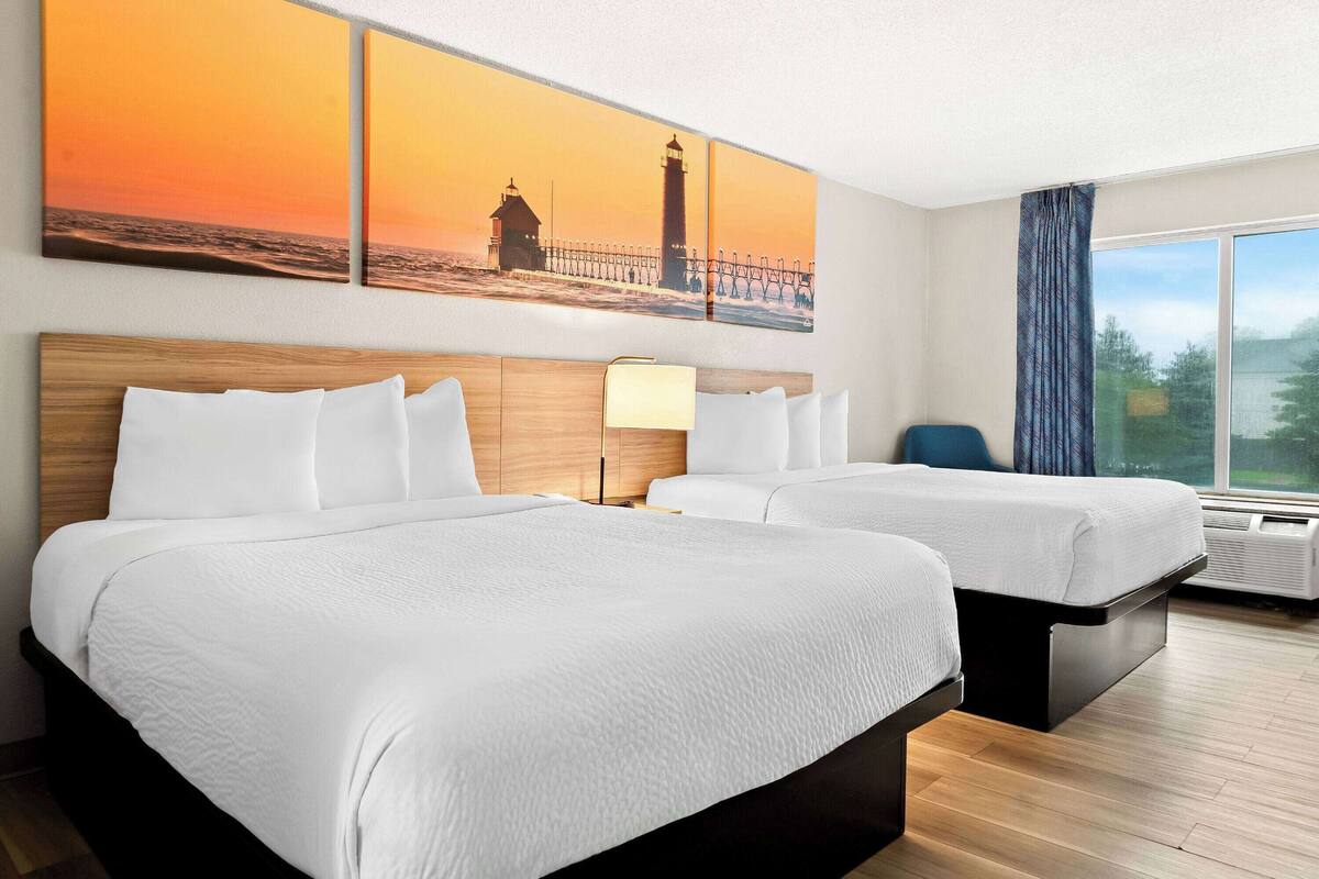 A hotel room with two beds, a large window with a view of a lighthouse, and a painting of a sunset over the ocean.