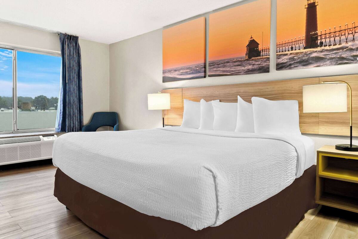 A hotel room with a large bed, a chair, a nightstand with a lamp, and a large window with a view of the sea and a lighthouse.