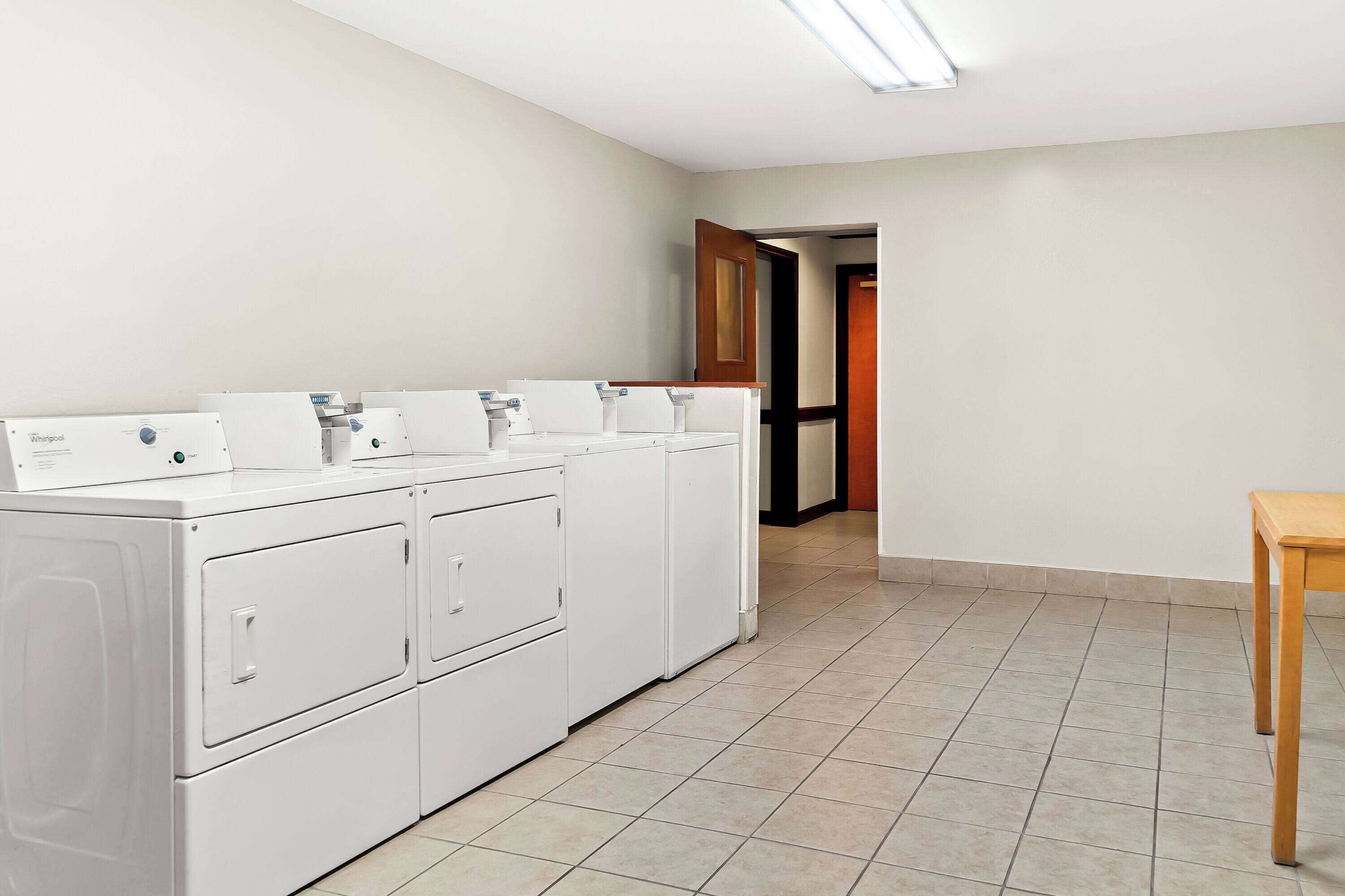 Un lavadero con una hilera de lavadoras y secadoras Whirlpool blancas, una mesa de madera y un piso de baldosas.