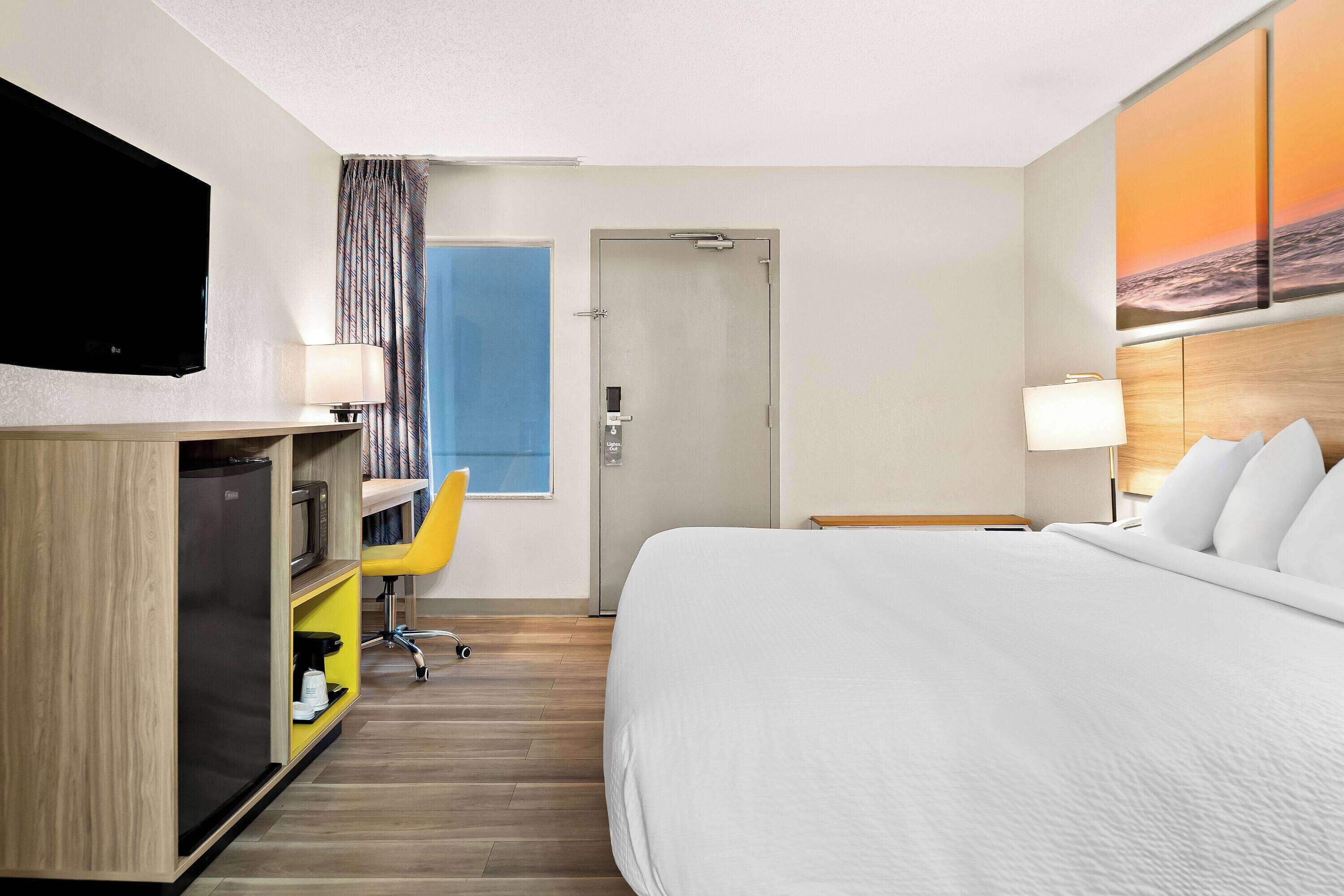 Una camera d'albergo con un letto, una scrivania con un computer, una TV, una finestra con tende e un quadro appeso al muro.
