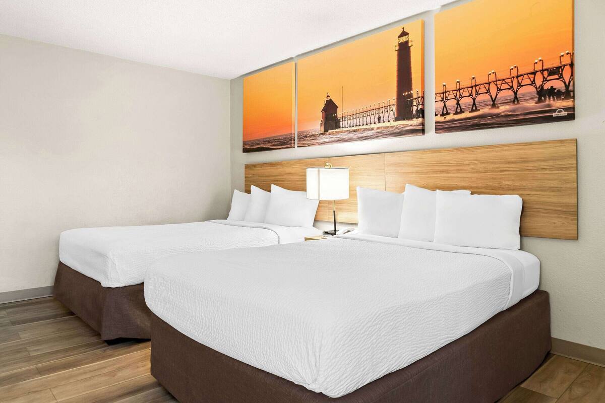 A hotel room with two beds, a wooden headboard, a nightstand with a lamp, and a large wall painting of a lighthouse and pier.