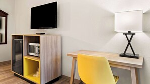 Ein Hotelzimmer mit Flachbildfernseher, Mikrowelle, Kühlschrank, Schreibtisch aus Holz, einem gelben Sessel und einer Nachttischlampe.